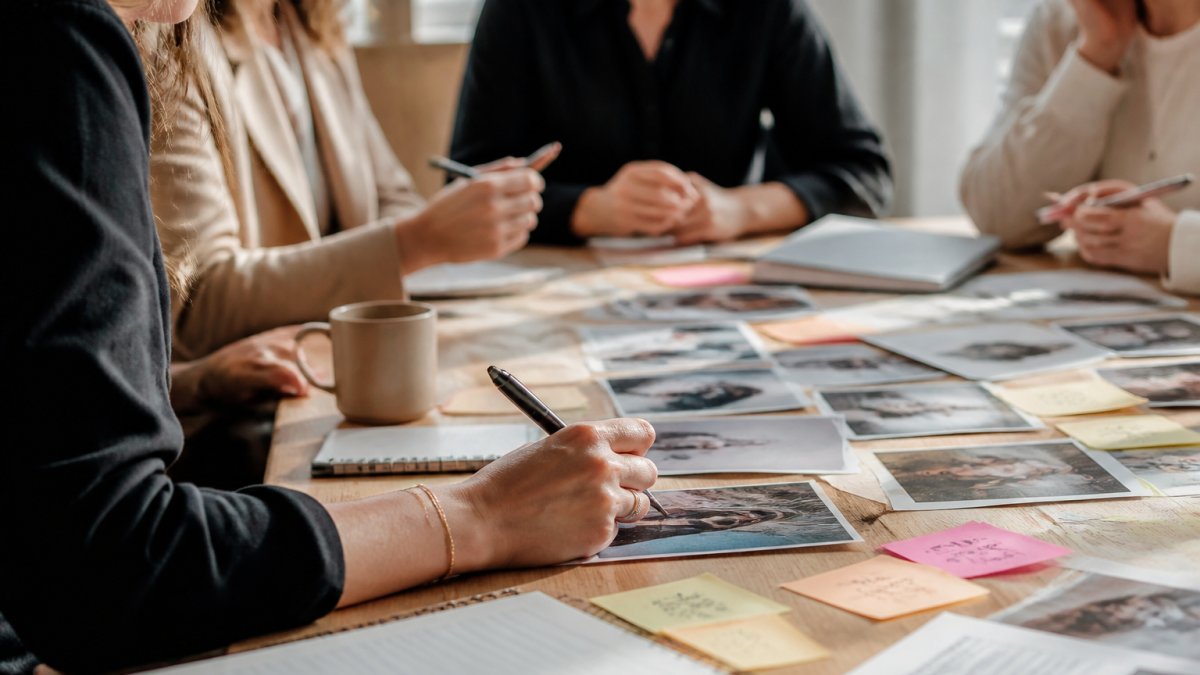 A diverse group of creative professionals gathered around a wooden table with notebooks and sticky notes, engaged in a brand vision workshop in a bright studio