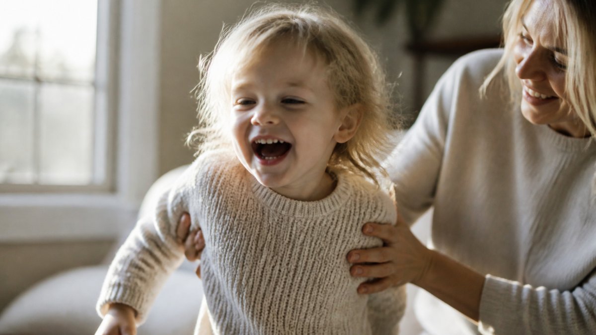 A young child in a simple knitted sweater laughing mid-movement in soft window light, with a parent's hands gently steadying them in the background