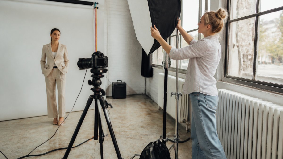 A fashion photographer adjusting a softbox on a studio set while a model in a styled outfit waits, a camera mounted on a tripod nearby and cables taped across the floor